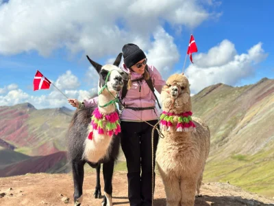 TourCompass-collega Iida staat op de top van de Rainbow Mountain in Peru, met twee vlaggen in de hand en een alpaca naast zich.
