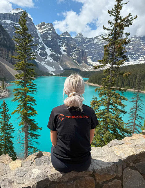 Een vrouw in een TourCompass-T-shirt kijkt uit over een turquoise meer en bergen met besneeuwde toppen in Canada